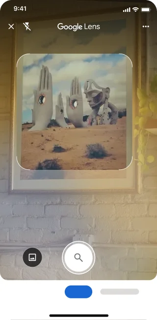 Sur un téléphone, l'application Google Lens est ouverte. Une notification indique à l'utilisateur qu'il peut appuyer sur le bouton de l'obturateur pour rechercher une image représentant de grandes sculptures de mains en pierre dans un désert.